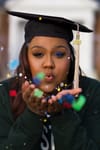 Image of a young Black woman 2021 graduate in cap and gown holding out hands and blowing confetti